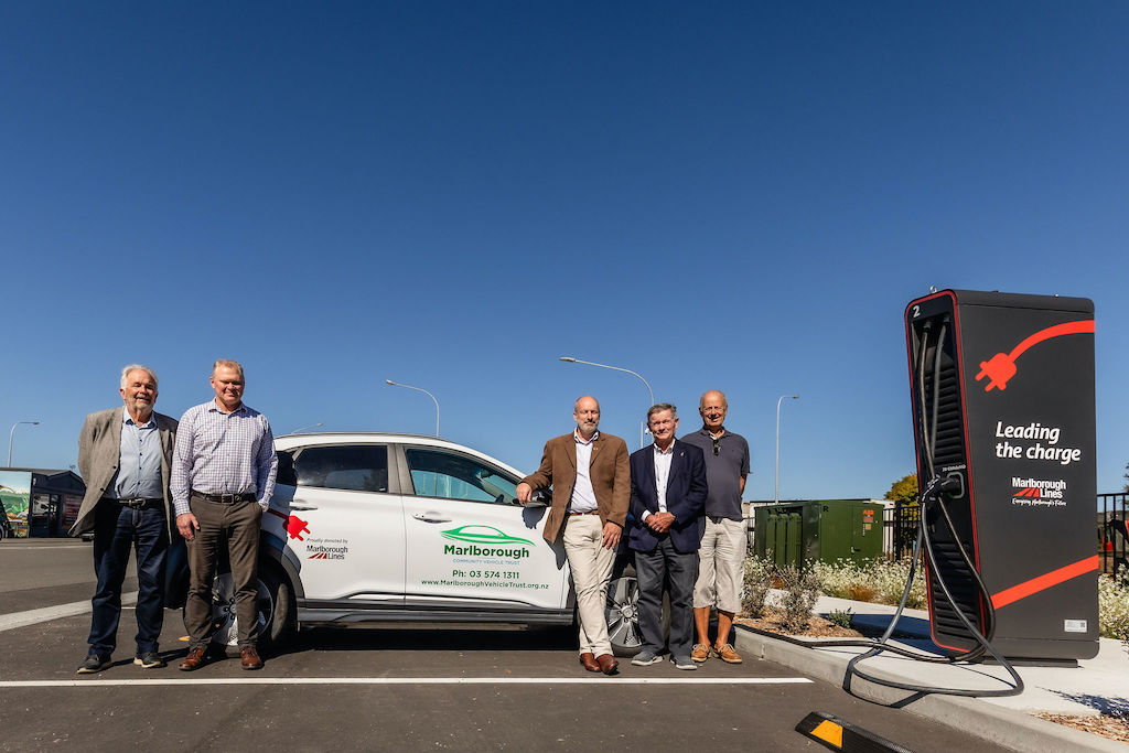 5 men stand in front of an electric vehicle parked at a charging station.